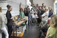 Raum mit einer Gruppe von Personen, links ein Tisch mit Schokoladenartikeln.
